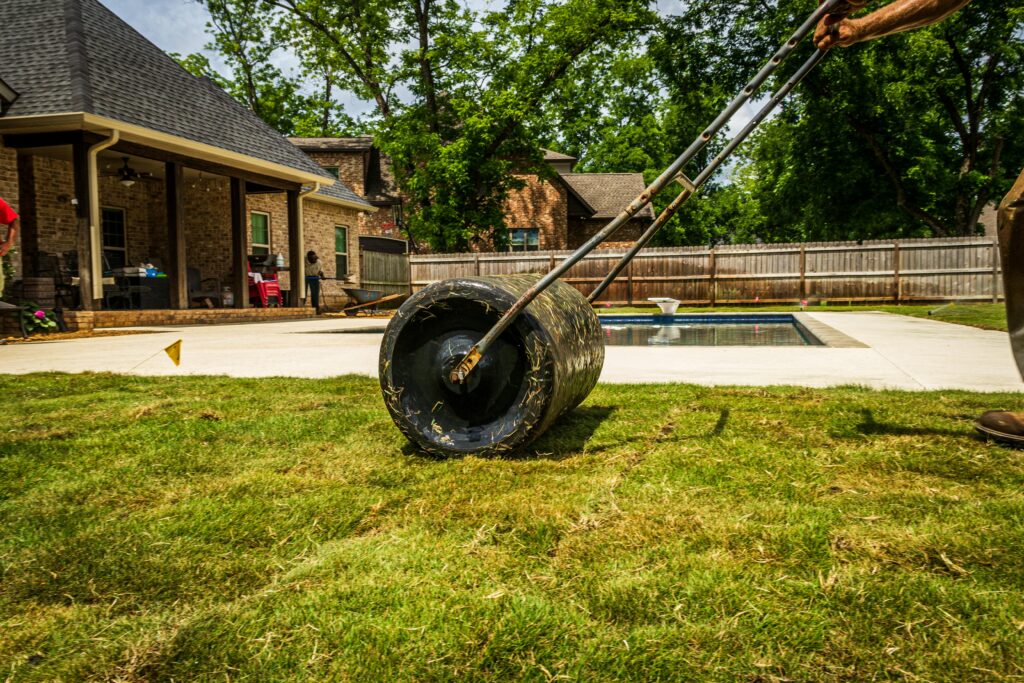 A worker rolling a lawn in a suburban backyard near a pool on a sunny day.